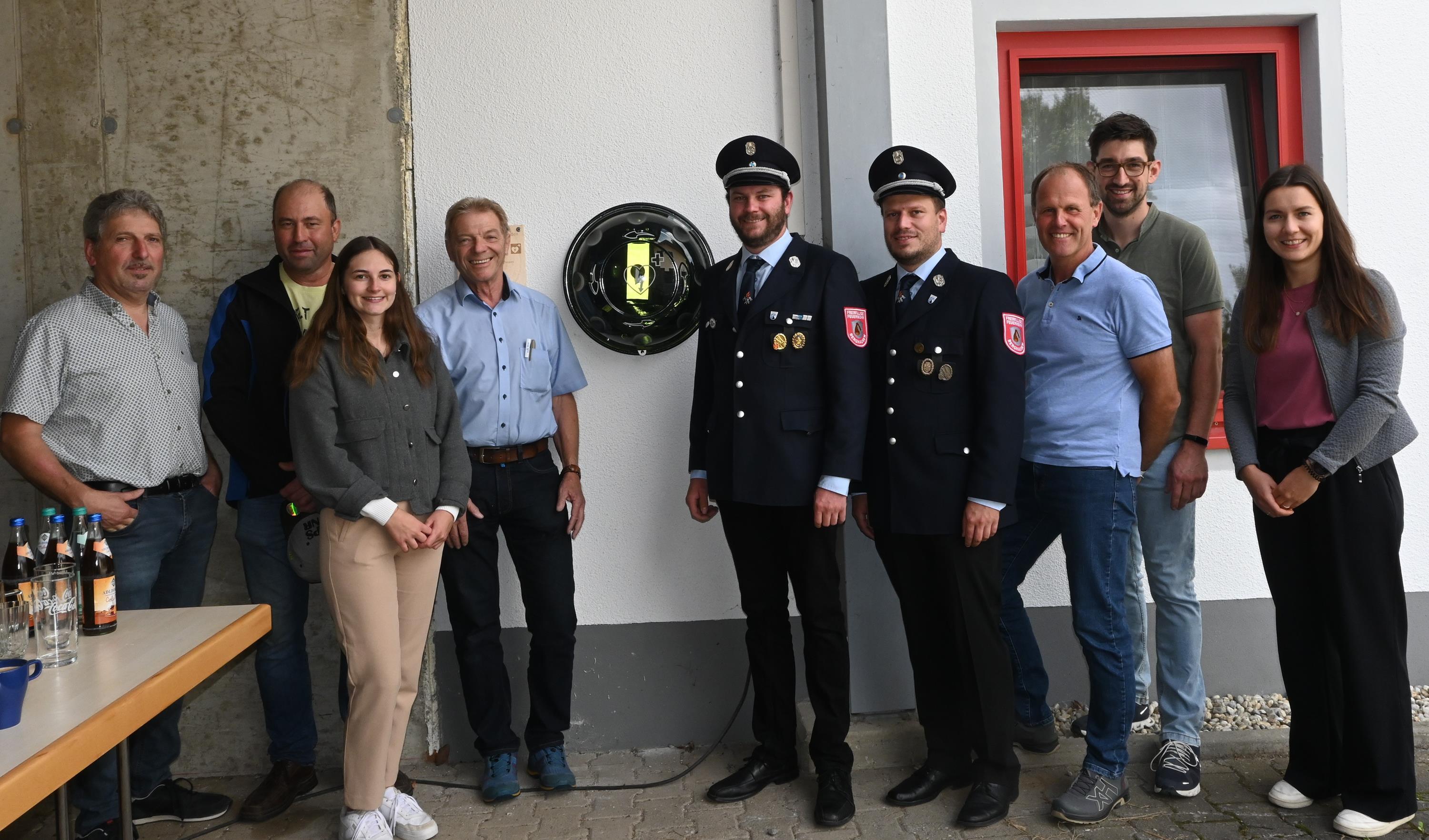 Neun Frauen und Männer flankieren den Defibrillator am Feuerwehrhaus