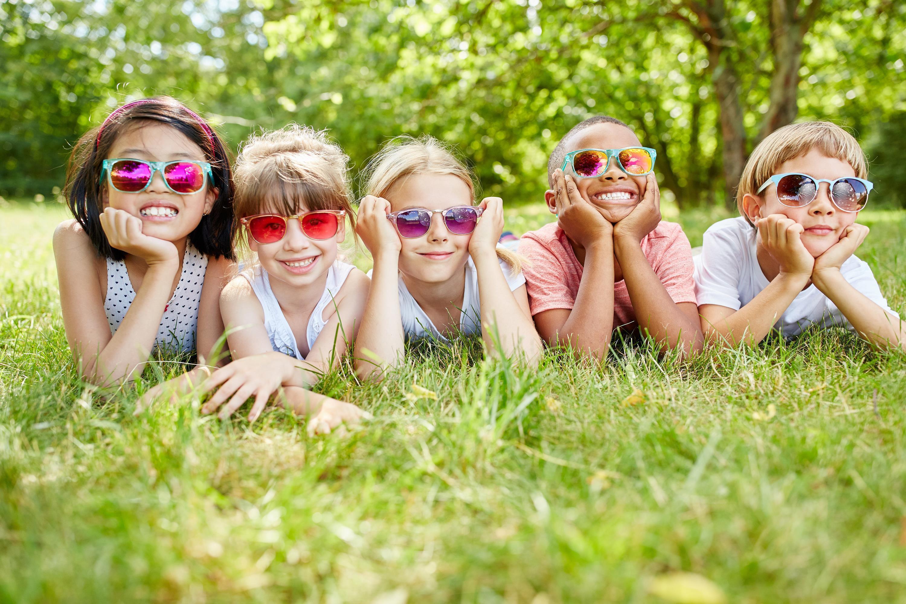 Fünf Kinder mit Sonnenbrillen liegen auf dem Bauch im Gras und lächeln