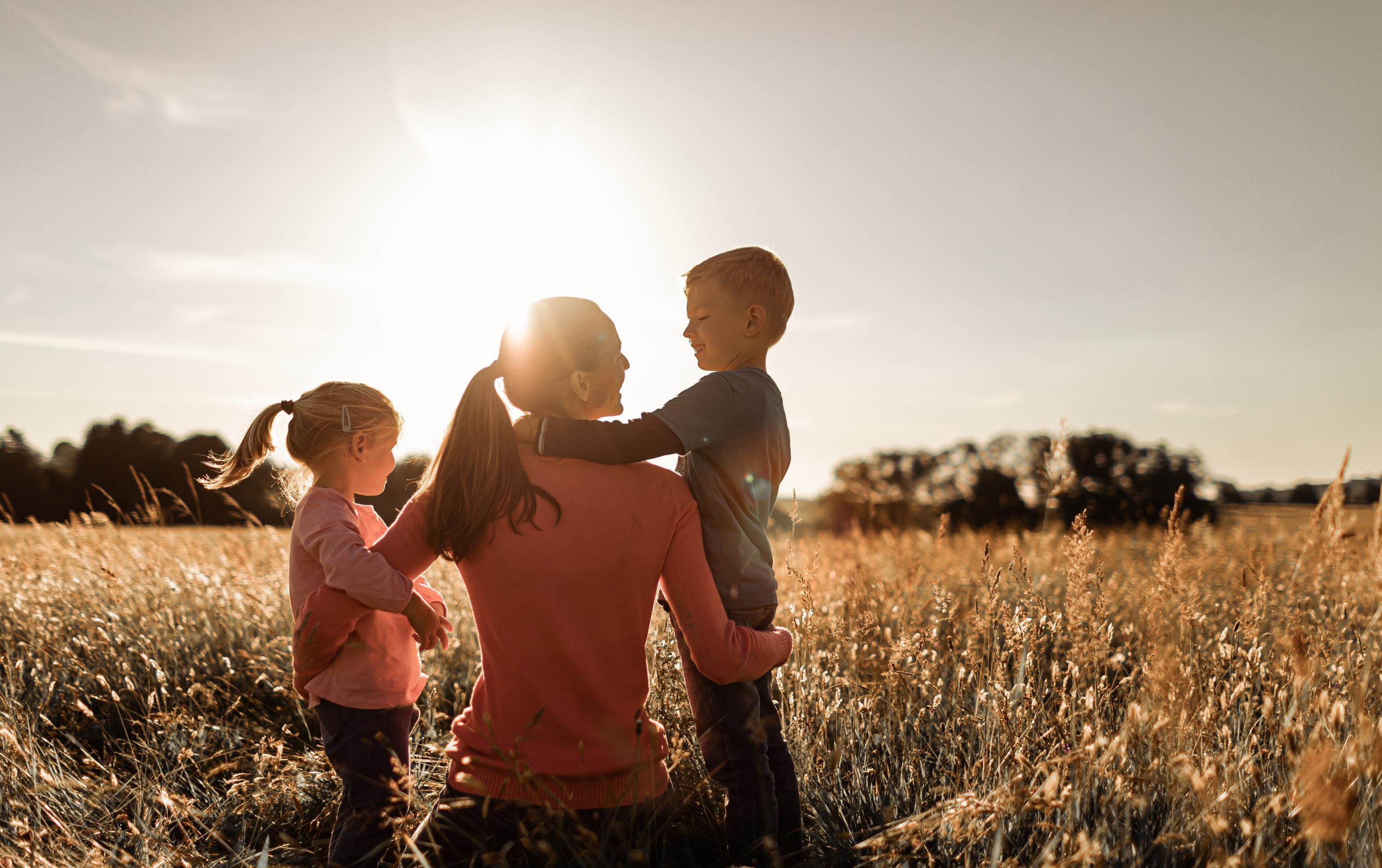 Mutter sitzt mit Tochter und Sohn im Arm in Wiese im Sonnenschein