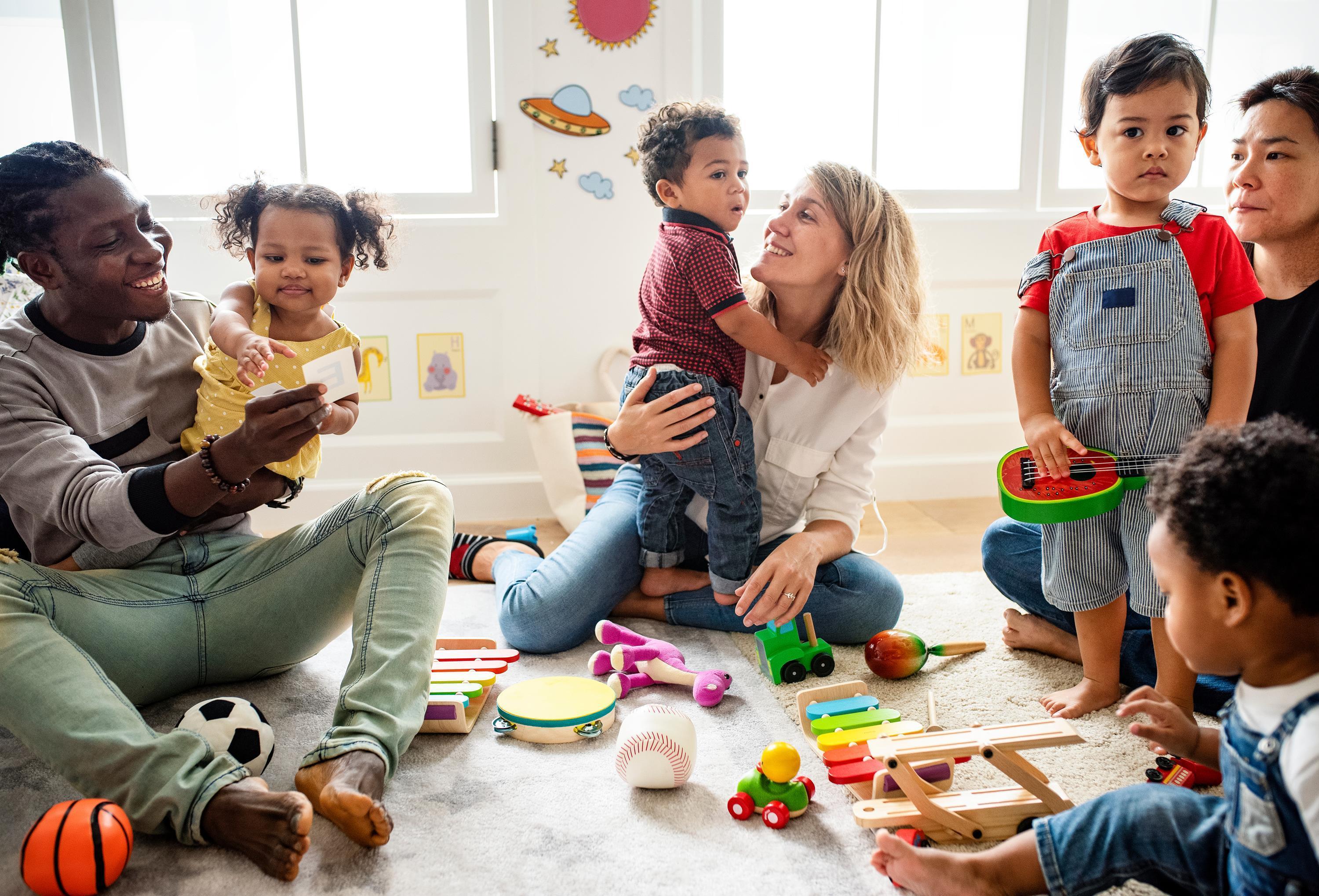 Vater und 2 Frauen sitzen am Boden wo vier Kinder miteinander spielen 