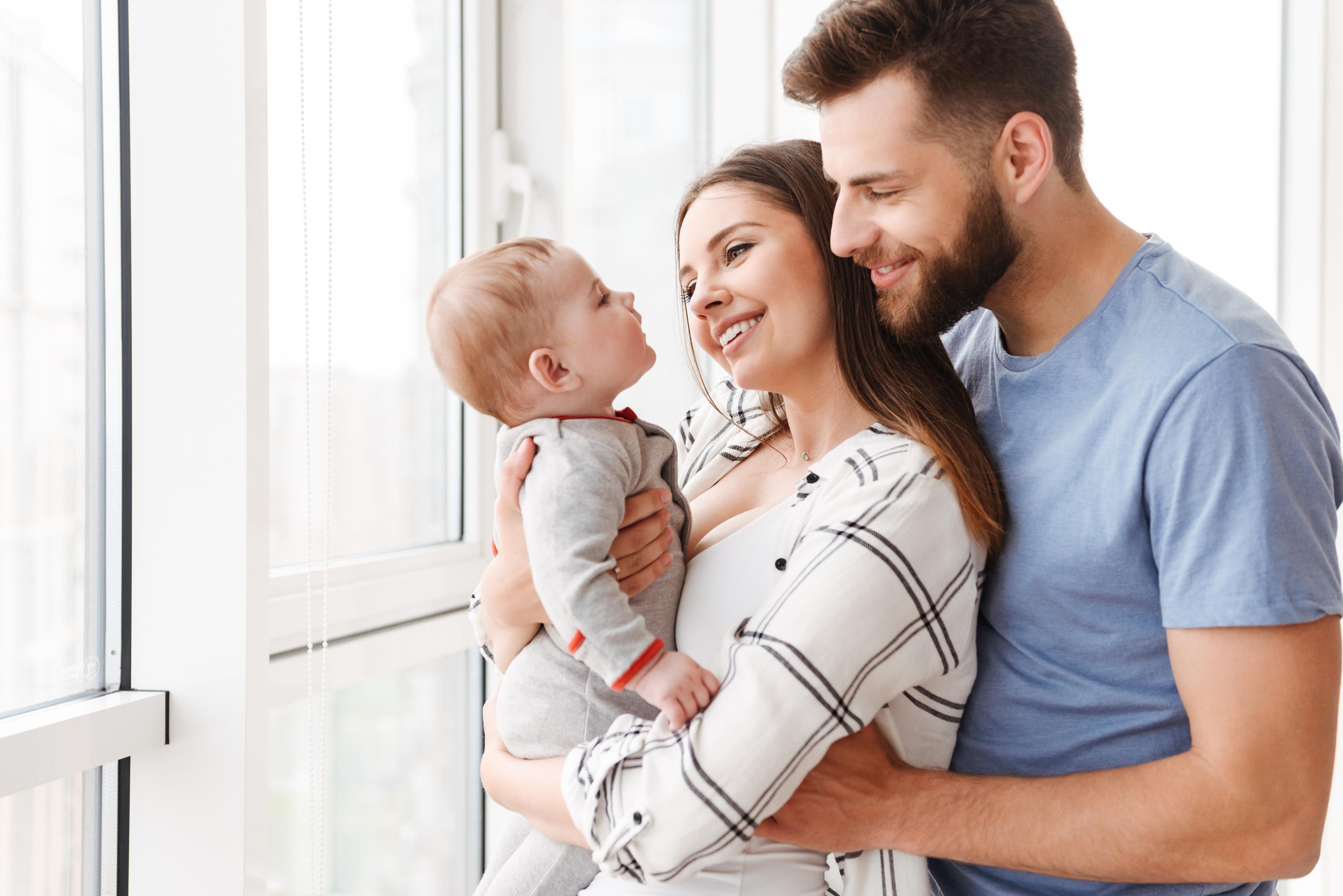Vater und Mutter, die ihr Baby auf dem Arm hält, stehen vor Fenster 