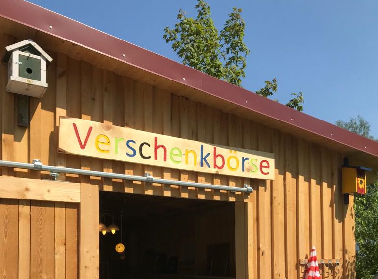 An einer großen Holzhütte hängt das Schild Verschenkbörse.