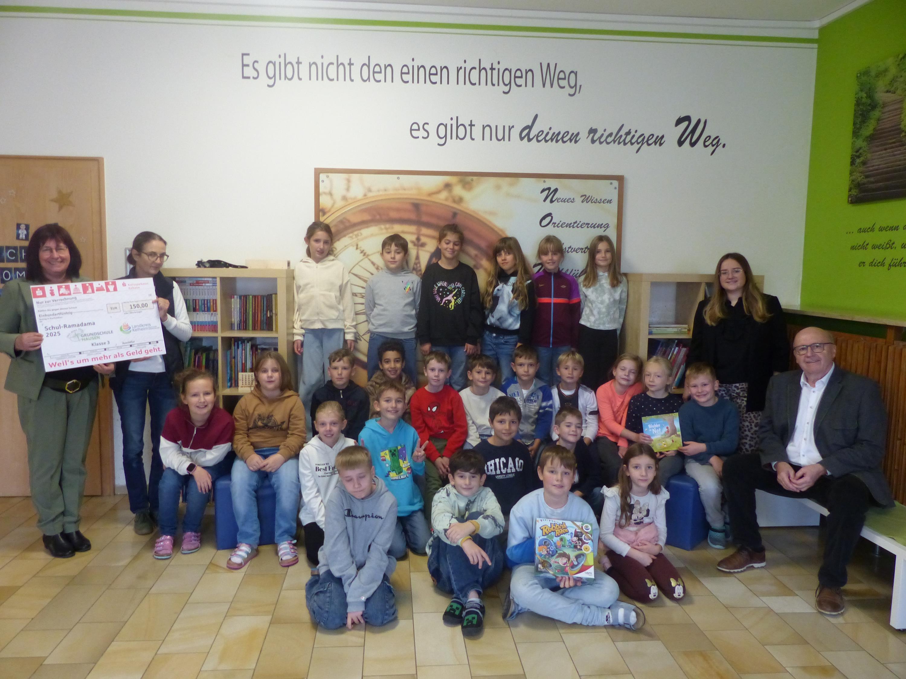 Eine Grundschulklasse lacht in die Kamera; Lehrerinnen halten Scheck