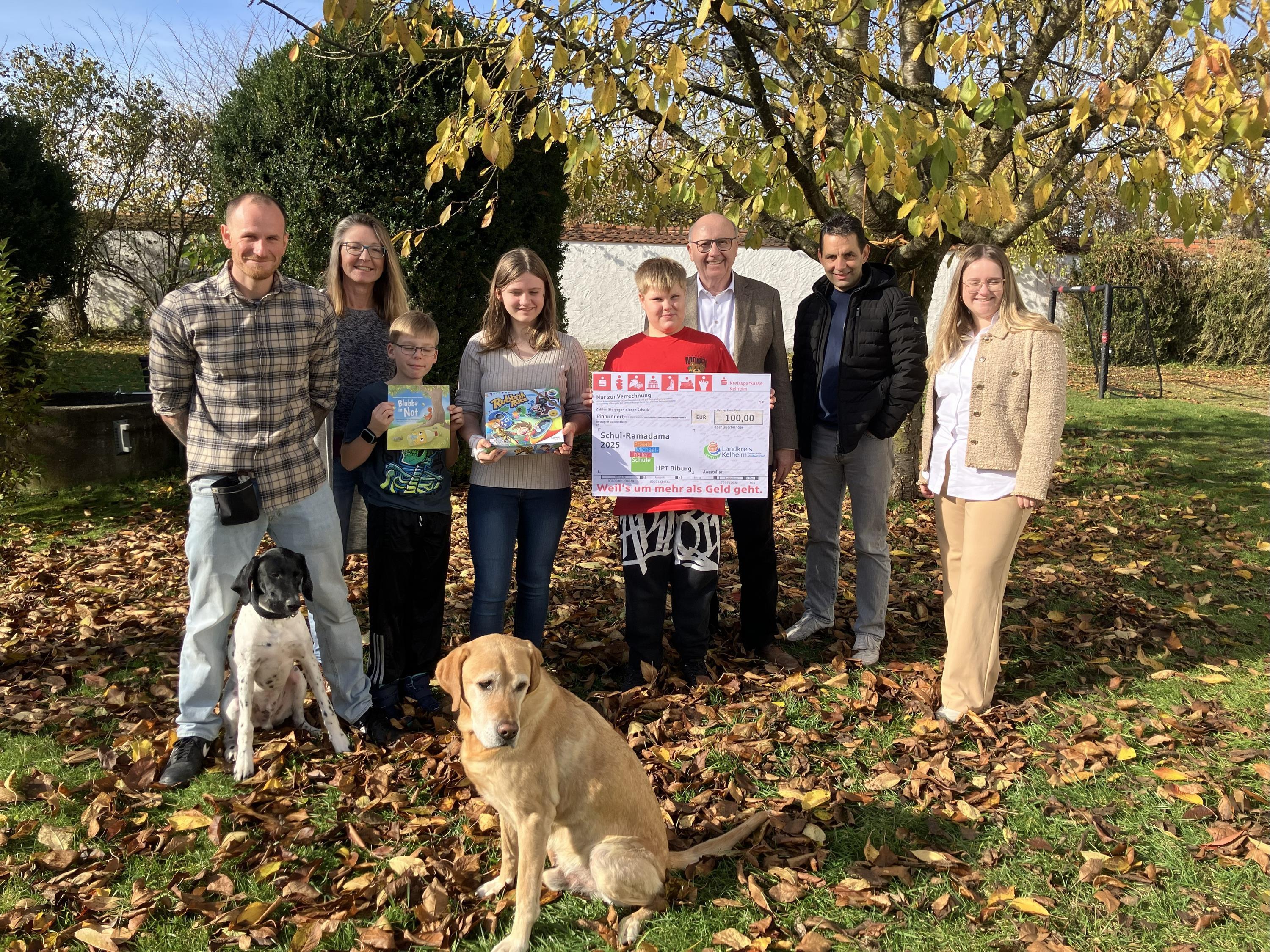 Acht Personen und zwei Hunde präsentieren Scheck unter einem Baum