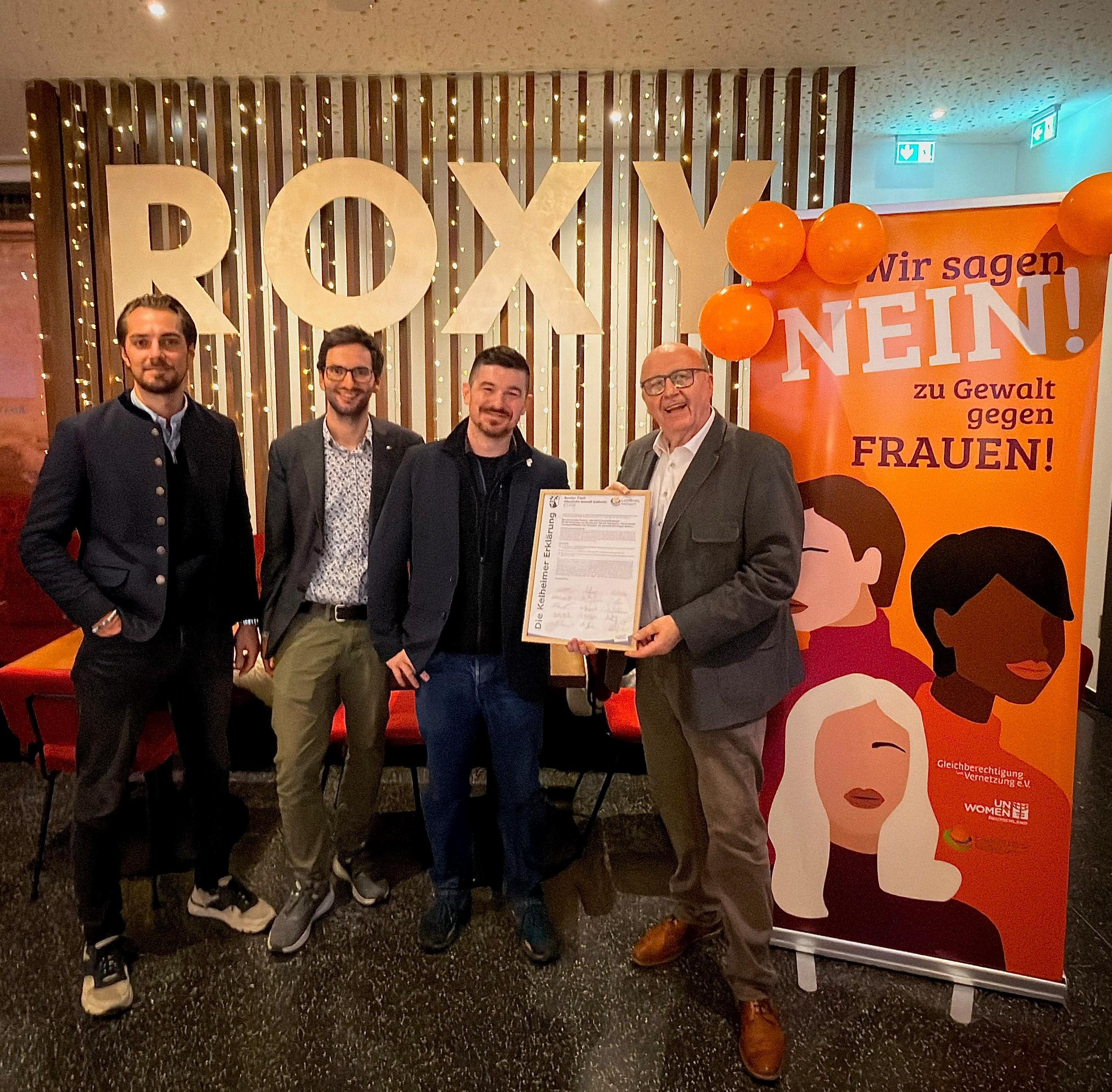 Vier Männer präsentieren im Kino-Foyer ein eingerahmtes Dokument.
