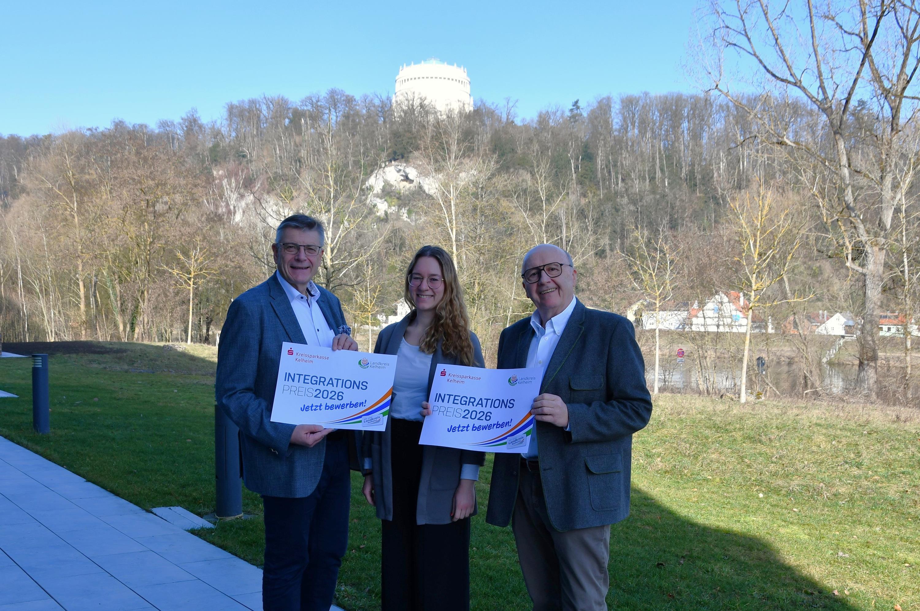 Drei Personen neben Landratsamt halten Integrationspreis-Plakat