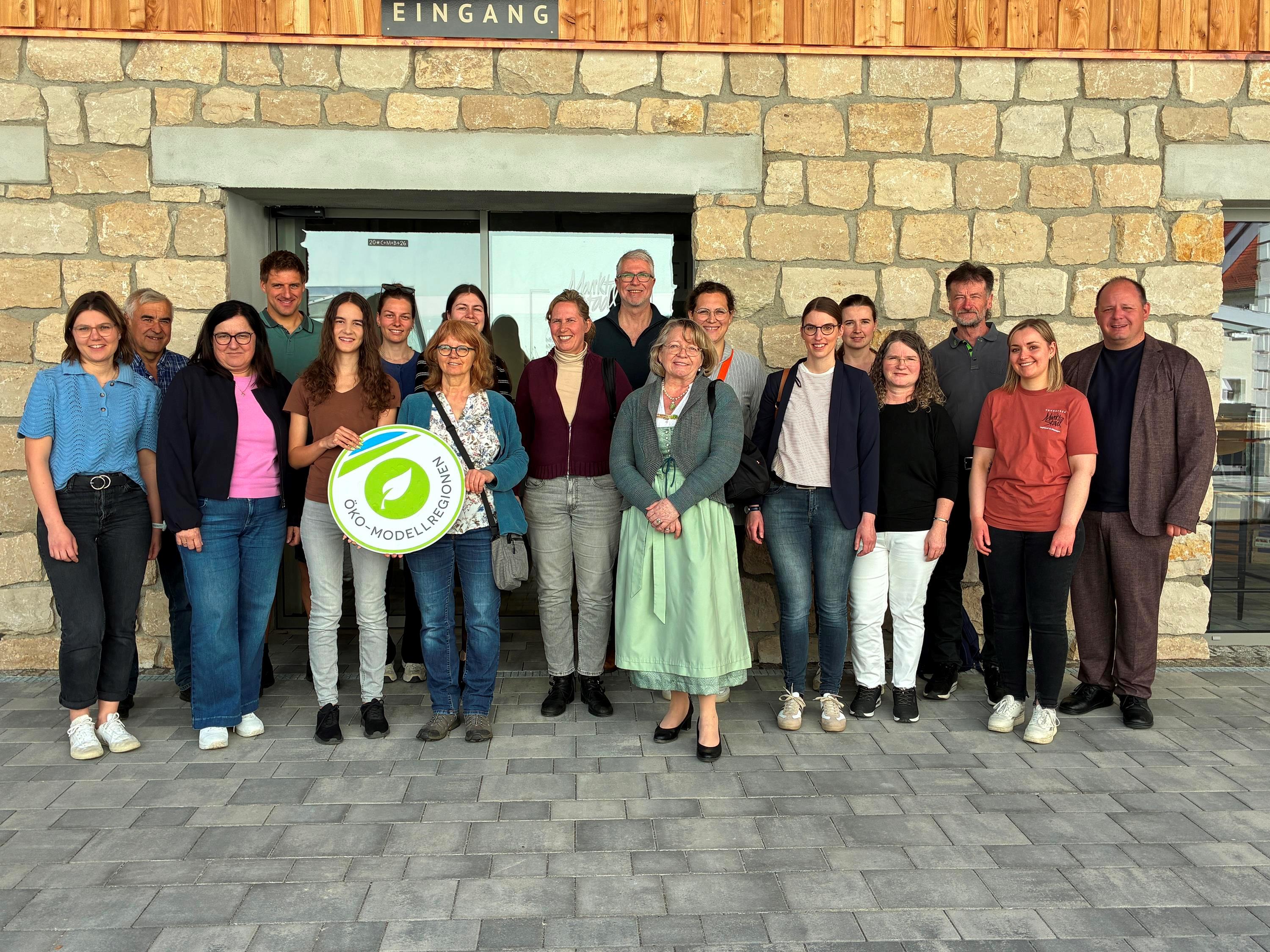 18 Personen vor einem umgebauten Stall, zwei halten ein rundes Schild