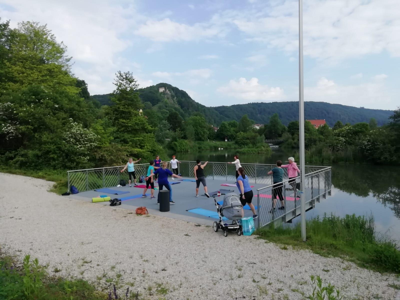 10 Frauen auf Bühne neben See machen Gymnastikbewegungen
