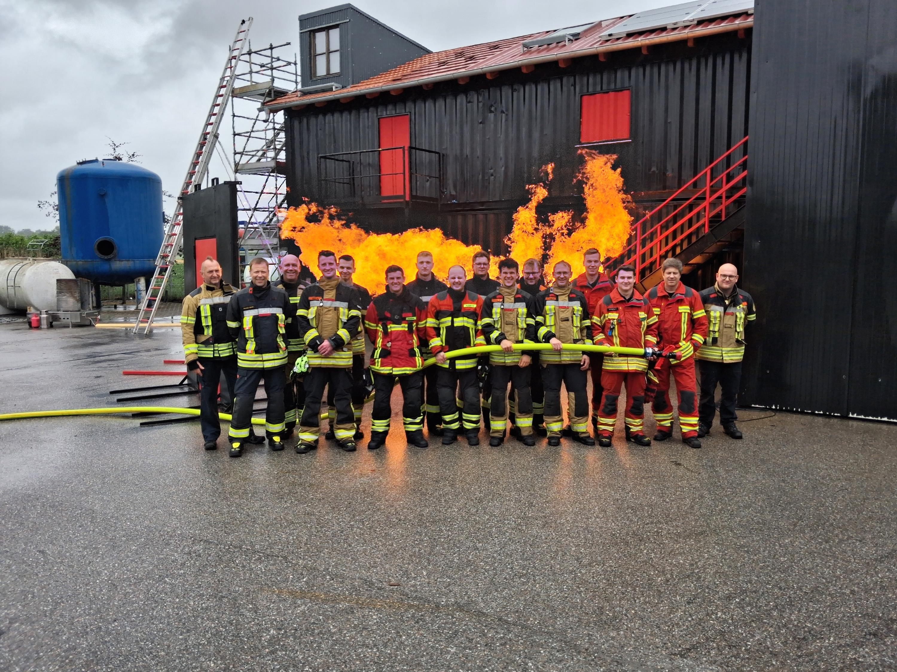 16 Feuerwehrmänner vor Flammen und einem schwarzen Container