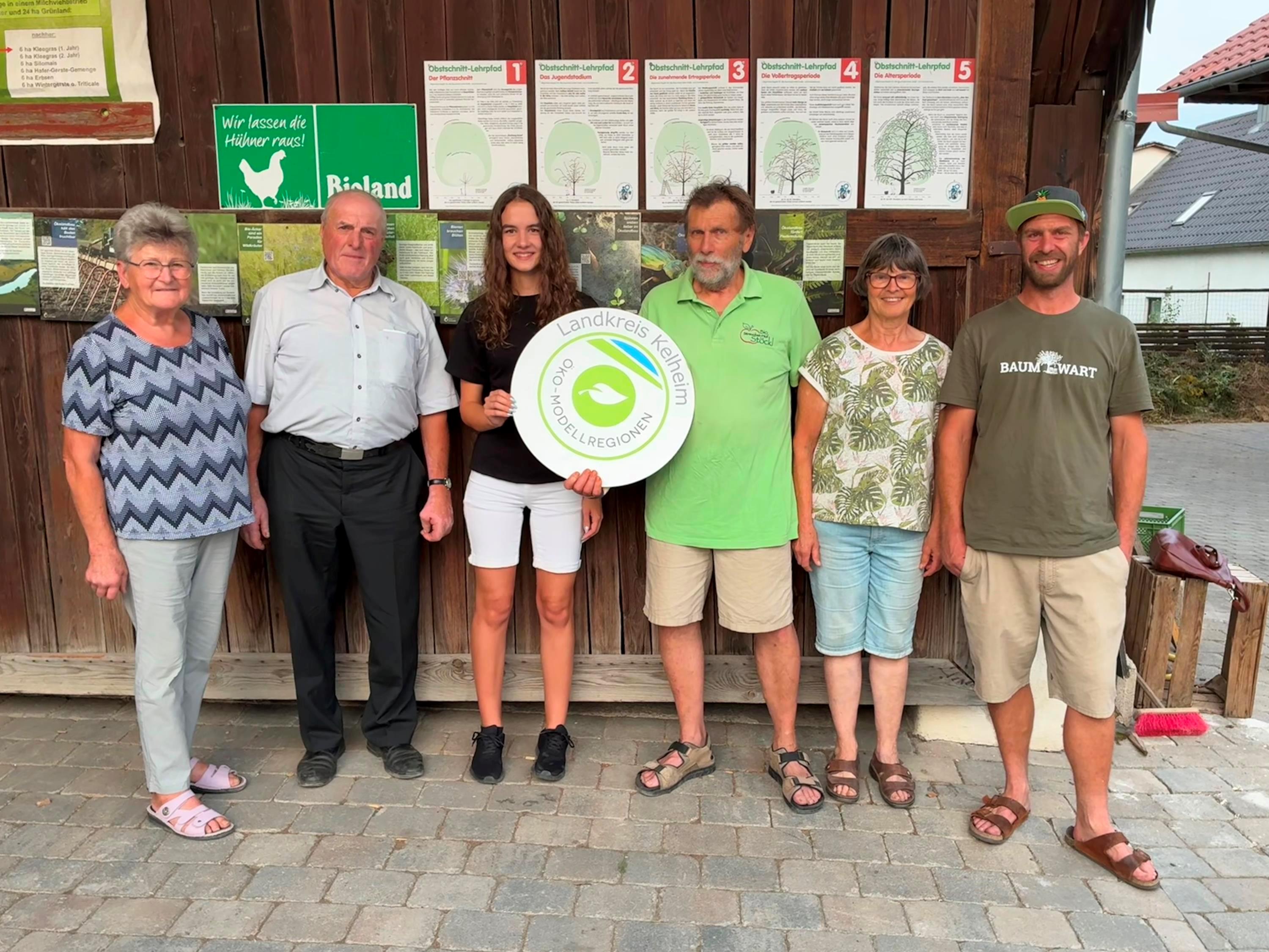 Frau mit rundem Schild in der Hand und fünf Personen vor Holzscheune