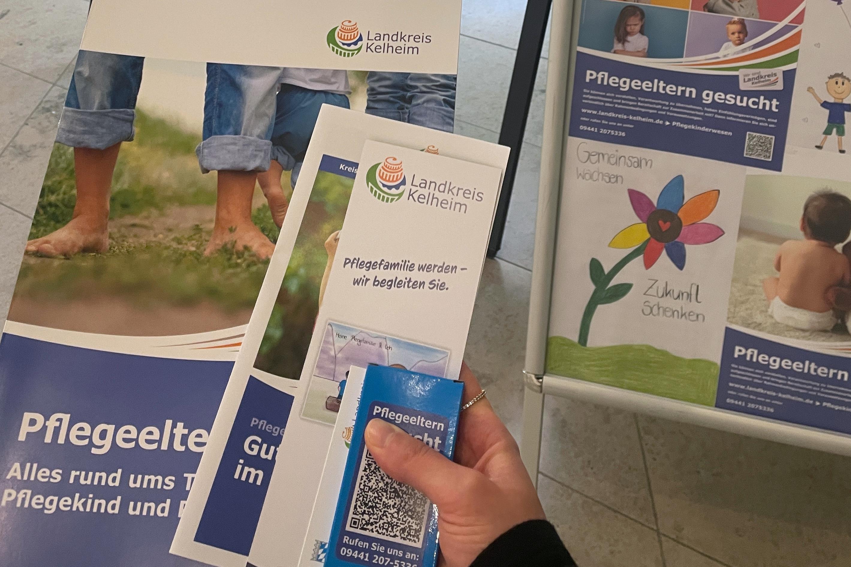 Eine Hand mit Infomaterialien vor einem Werbeaufsteller.